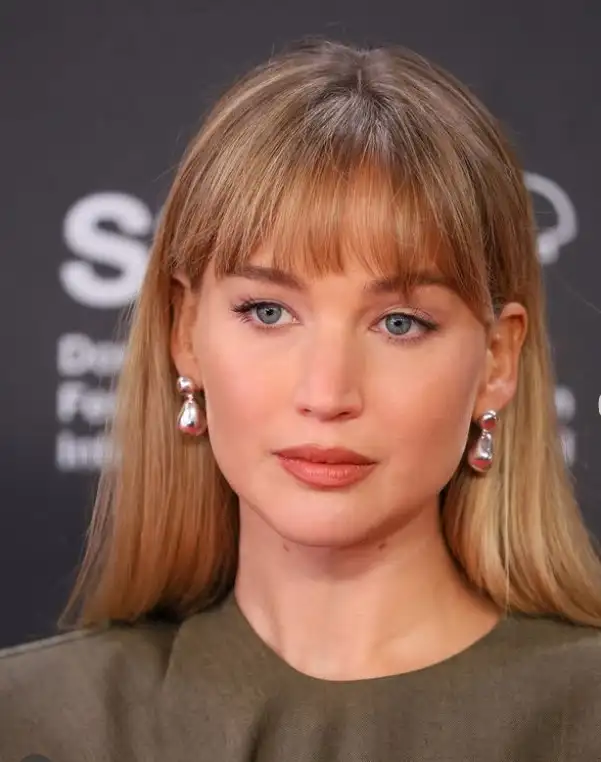 Jennifer Lawrence layered bangs with soft volume, blending naturally into medium-length hair for a fuller, textured hairstyle.