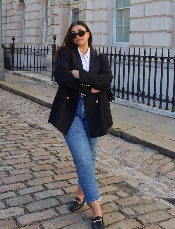 Woman wearing dark blue jeans with a structured black blazer, light top, and black loafers.