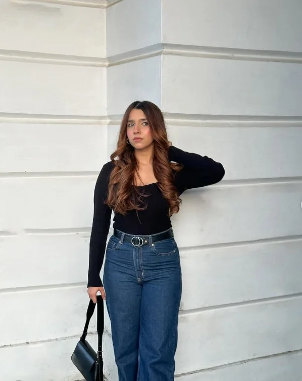 Woman styling dark blue jeans with a fitted black long-sleeve top, gold jewelry, and black pointed shoes.