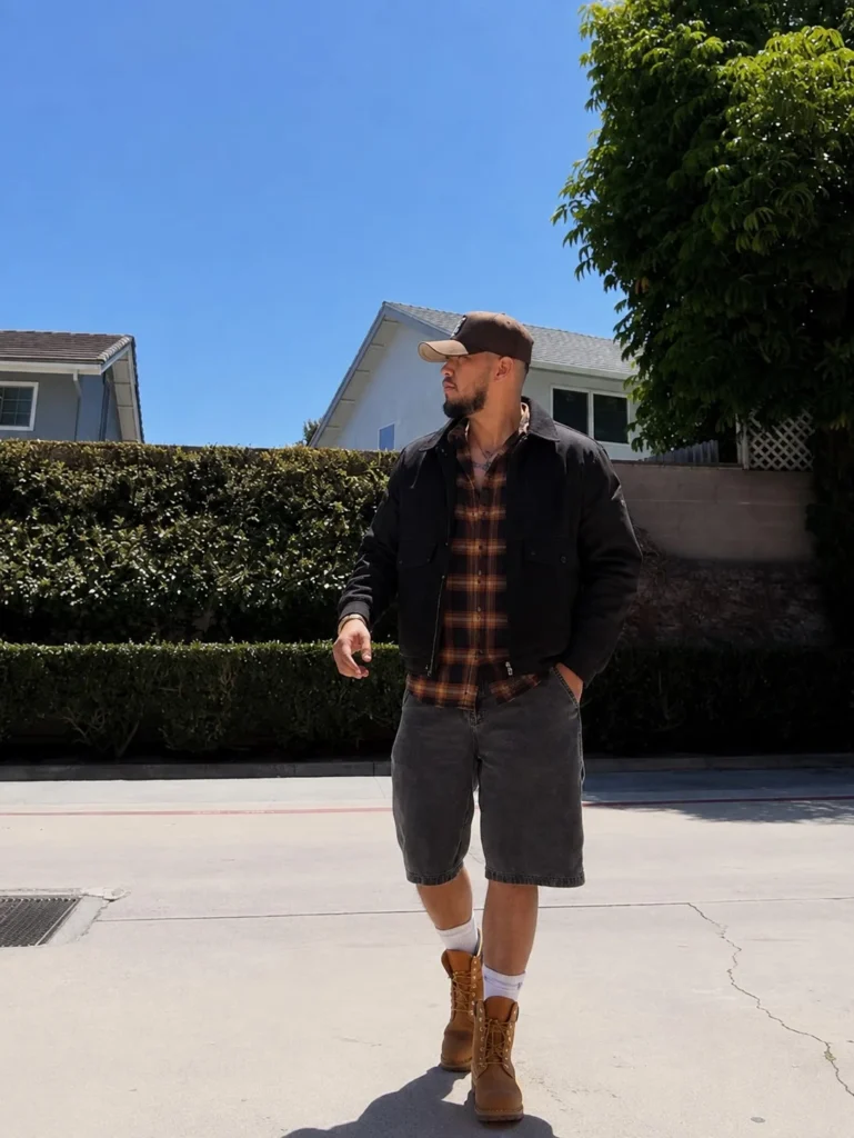 Man wearing a brown flannel shirt layered under a black jacket with dark jeans and casual boots.