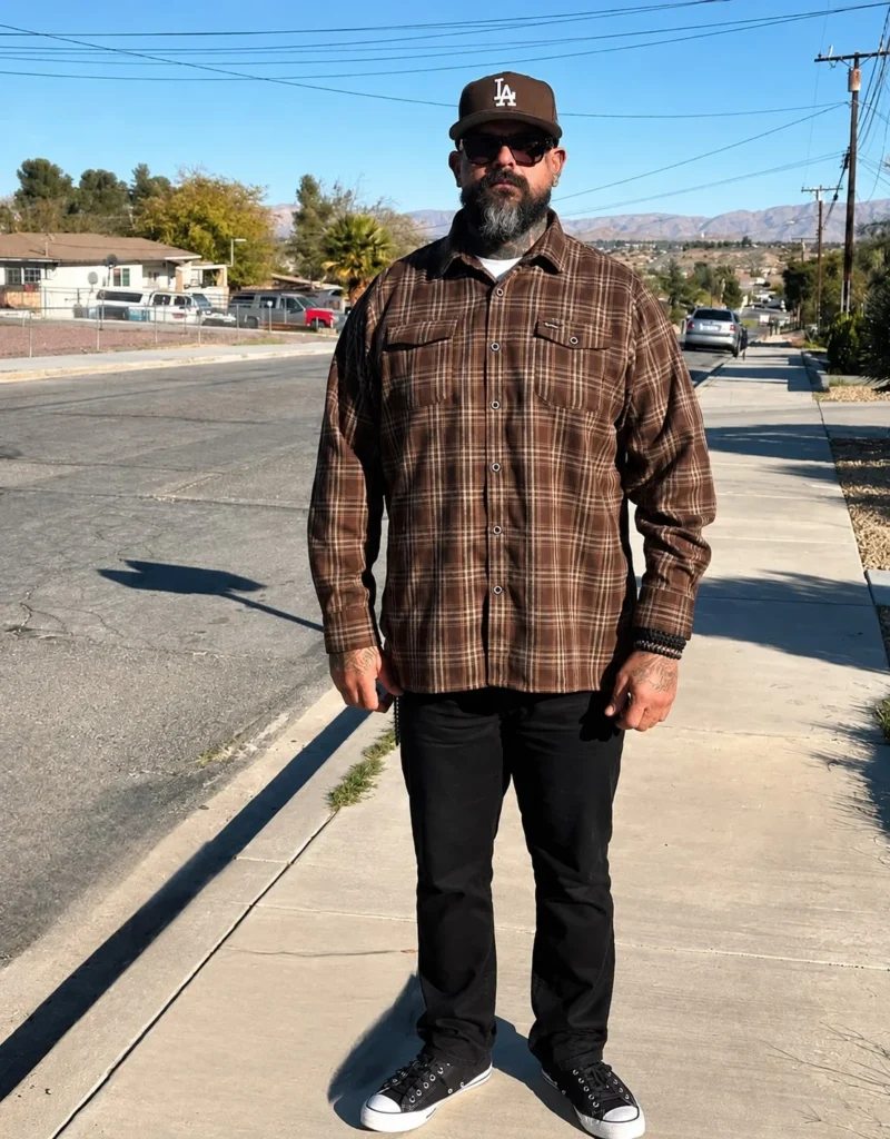 Man wearing a brown plaid flannel shirt over a cream T-shirt with black jeans and black leather boots.