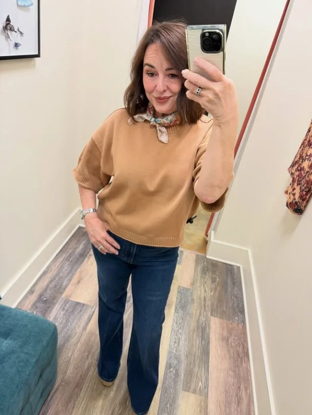 Woman wearing dark blue jeans with a soft camel sweater, brown loafers, leather belt, and structured handbag.