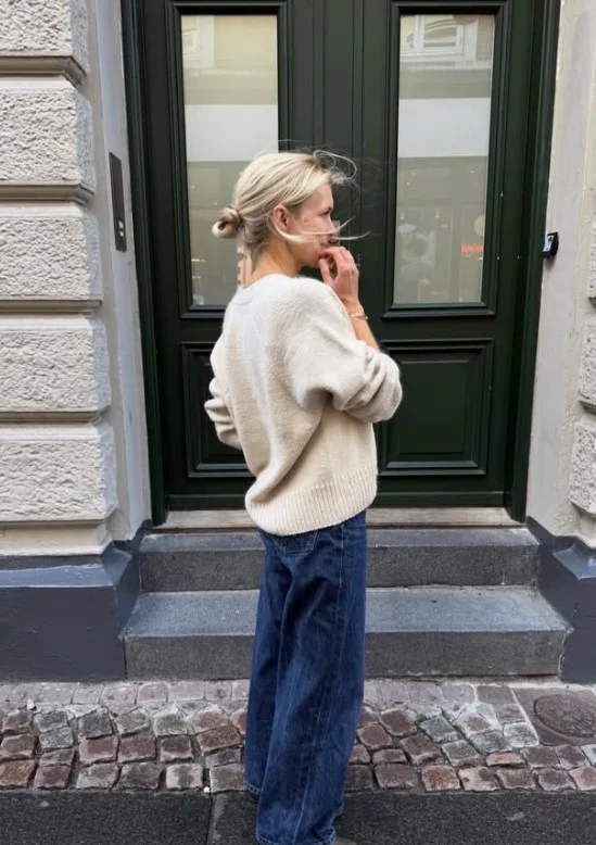 Woman wearing dark blue jeans with a cream cardigan, brown loafers, and a simple shoulder bag.