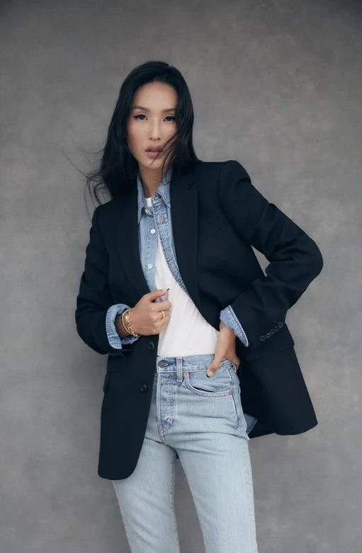 Woman wearing a blue denim button-up shirt under a black blazer with black trousers.
