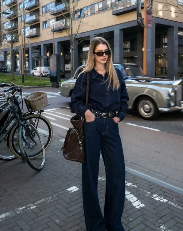 Woman wearing a blue denim button-up shirt with dark indigo jeans and a sculptural belt.