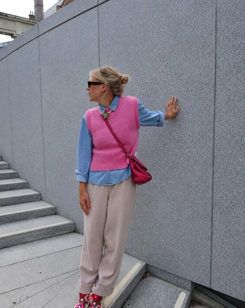 Woman wearing a blue denim button-up shirt under a cream knit vest with dark trousers.