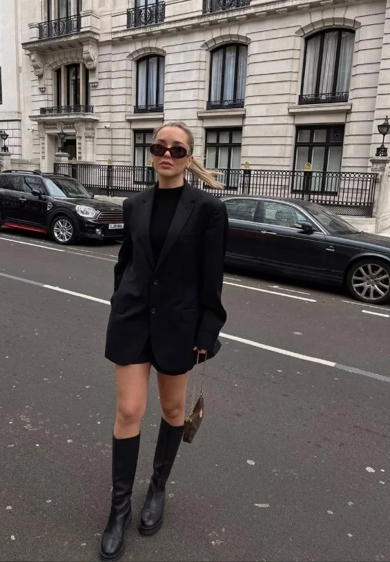 Woman wearing long black boots with a black mini dress, opaque tights, and a camel coat.