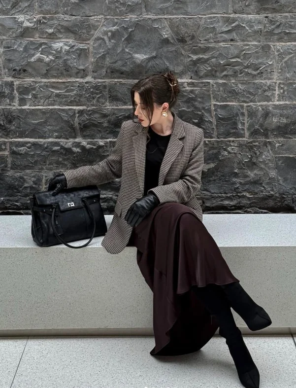 Woman wearing long black boots with a satin midi skirt, cardigan, and structured coat.