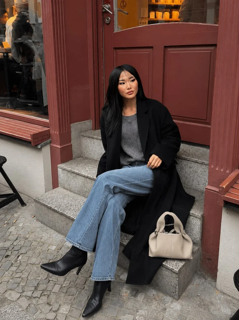 Woman wearing straight-leg jeans, a neutral knit, long black boots, and a long coat