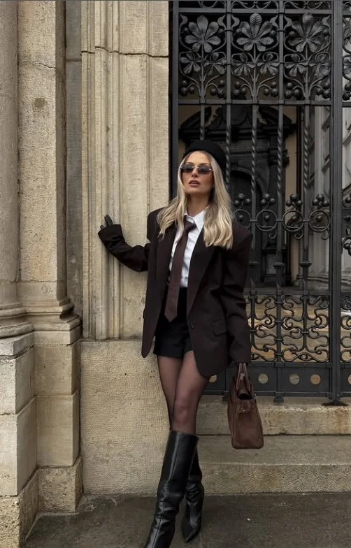 Woman wearing long black boots with a white button-down shirt, black blazer, and tailored skirt.