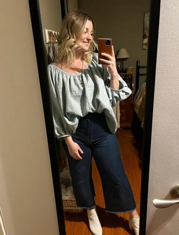 Woman wearing dark blue jeans with an olive green top, brown leather accessories, and ankle boots.