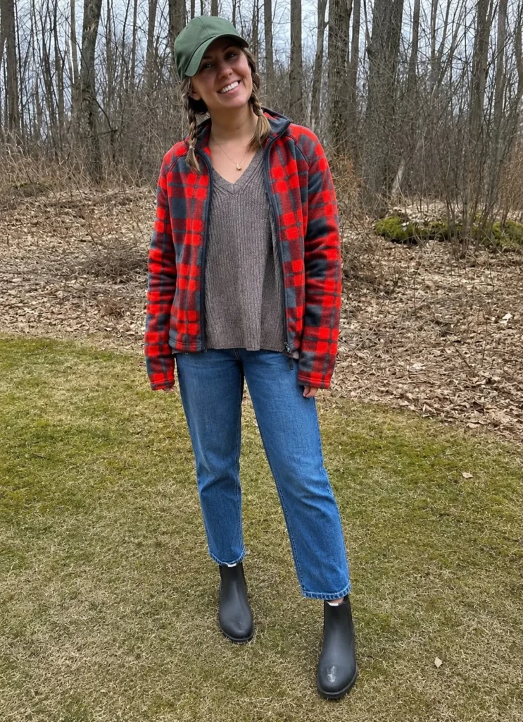 Woman wearing short black rain boots with straight-leg blue jeans, a cream sweater, and a trench coat.