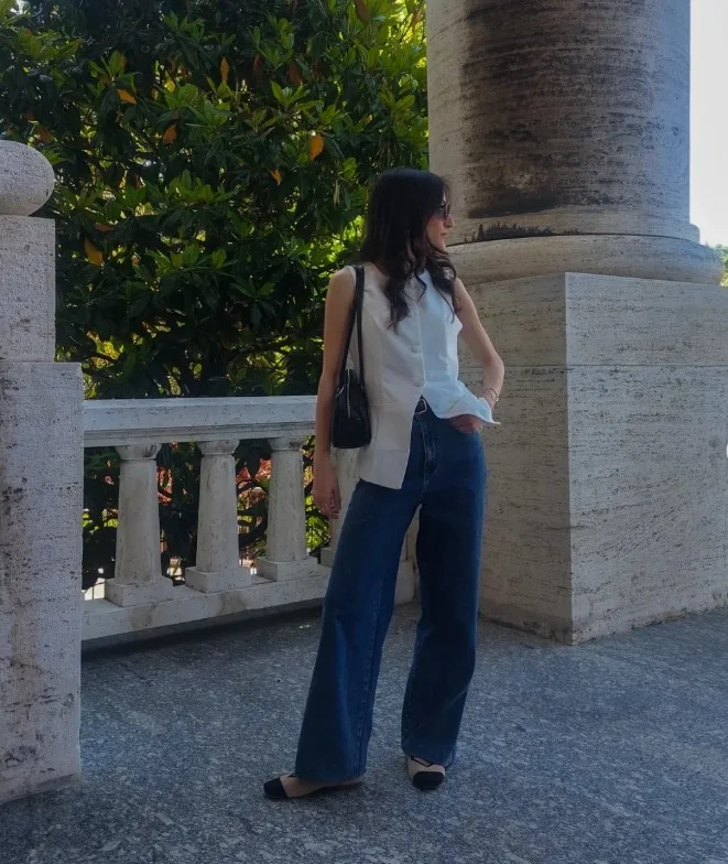 Woman wearing dark blue jeans with a tailored cream vest over a fitted top and polished loafers.