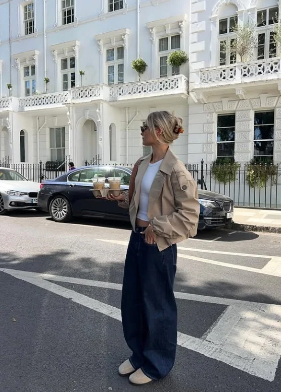 Woman wearing dark blue straight-leg jeans with a beige trench coat, simple white top, and loafers.