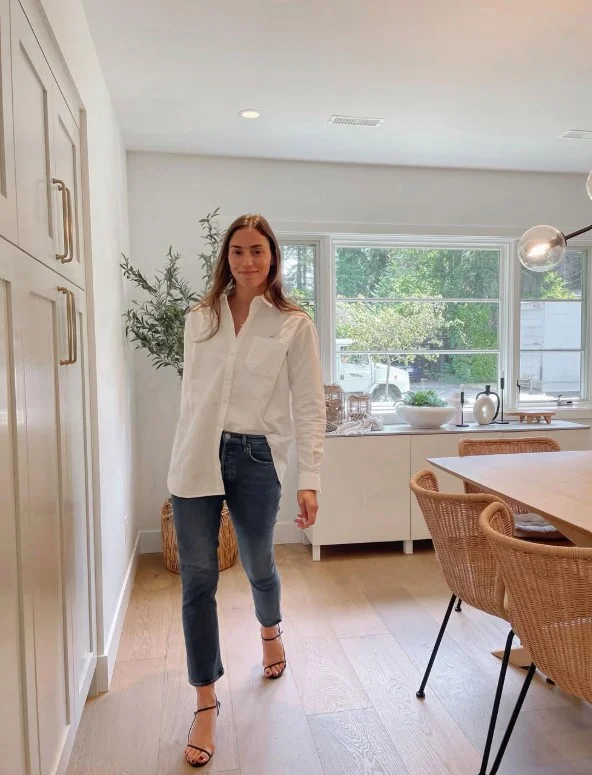 Woman wearing dark blue straight-leg jeans with a crisp white button-down shirt, brown leather belt, and loafers.