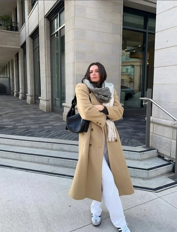 Woman wearing white jeans with a camel coat, neutral sweater, and brown boots for a sophisticated cold-weather outfit.
