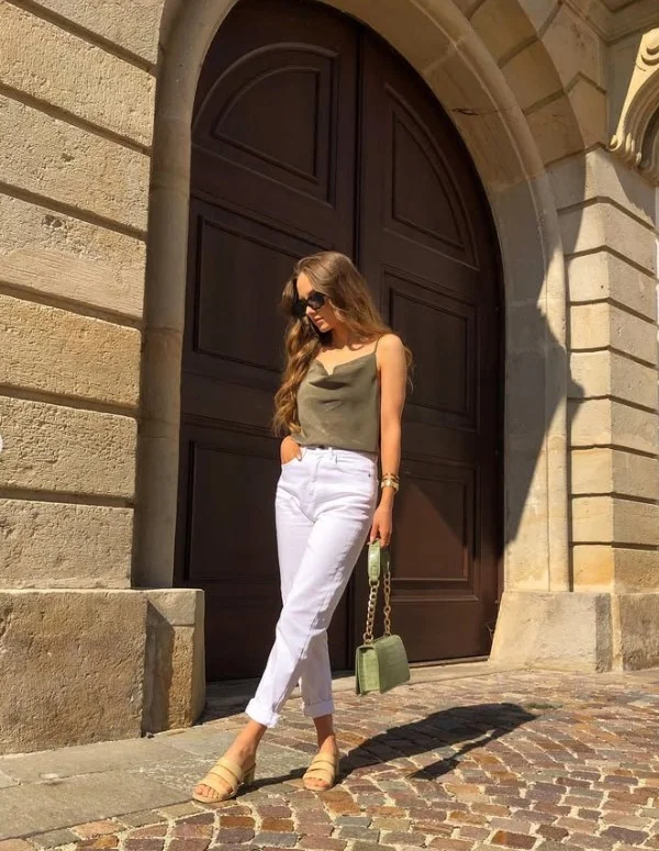 Woman wearing white jeans with a simple strappy top, flat sandals, sunglasses, and a woven bag for a relaxed summer outfit.