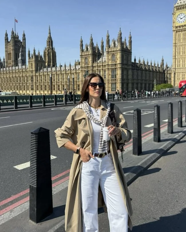 Woman wearing white jeans with a classic trench coat, simple top, and loafers for a polished transitional outfit.