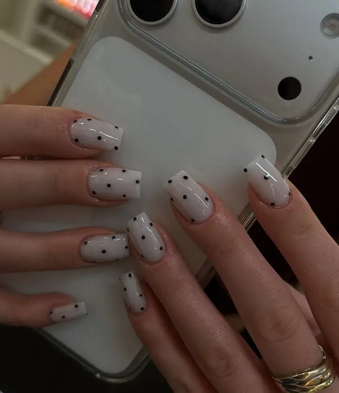 White acrylic nails decorated with evenly spaced black polka dots.